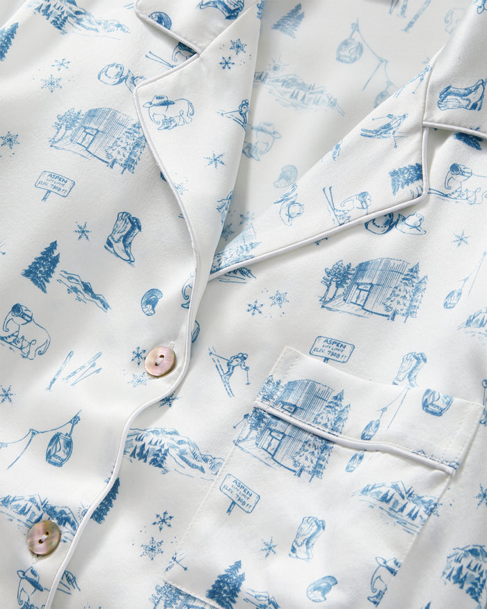 Petite Plume x White Elephant Women’s Silk Pajama Set features a white pajama top adorned with blue winter-themed illustrations—including cabins, pine trees, bears, skiing scenes, and snowflakes—and is detailed with white piping, buttons, and a chest pocket.