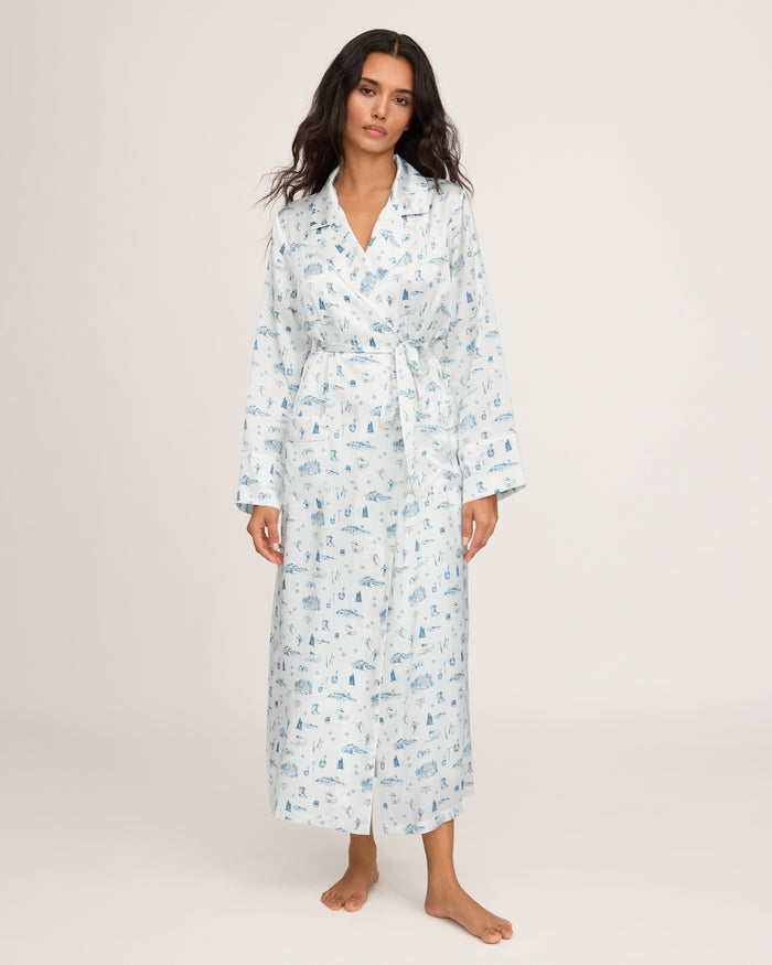 A woman stands barefoot against a plain, light background, wearing the Petite Plume x White Elephant Women’s Silk Robe—a long, light blue and white printed robe with a belt.