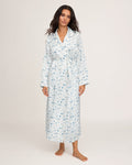 A woman stands barefoot against a plain, light background, wearing the Petite Plume x White Elephant Women’s Silk Robe—a long, light blue and white printed robe with a belt.