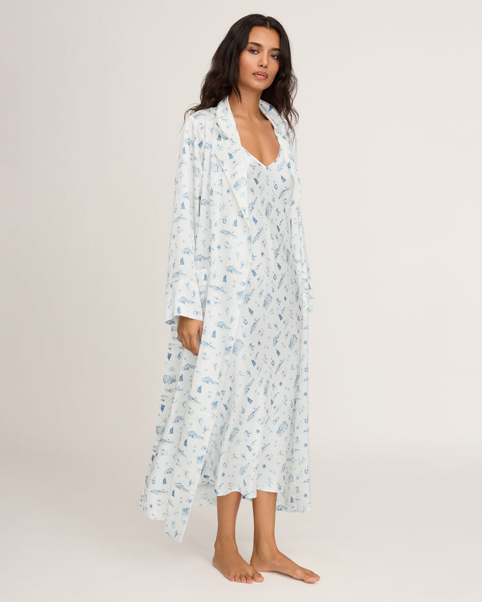 A woman with long dark hair poses barefoot against a plain, light background wearing the Petite Plume x White Elephant Women’s Silk Robe—a long, light blue patterned robe paired with a matching nightgown.