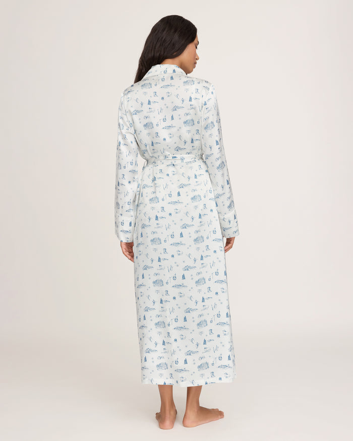 A person stands barefoot facing away, wearing the Petite Plume x White Elephant Women’s Silk Robe—a long white silk robe adorned with light blue patterns. The robe reaches their ankles, features long sleeves, a belted waist, and is set against a plain off-white background.