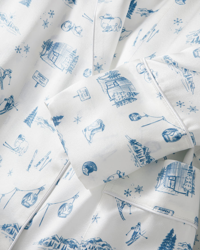 The Petite Plume x White Elephant Women’s Silk Robe features a white fabric adorned with blue sketches of ski cabins, skiers, ski gear, and snowy mountain scenes. Accented by shirt cuffs and buttons, the robe showcases repeating winter sports and alpine nature motifs throughout its luxurious silk design.