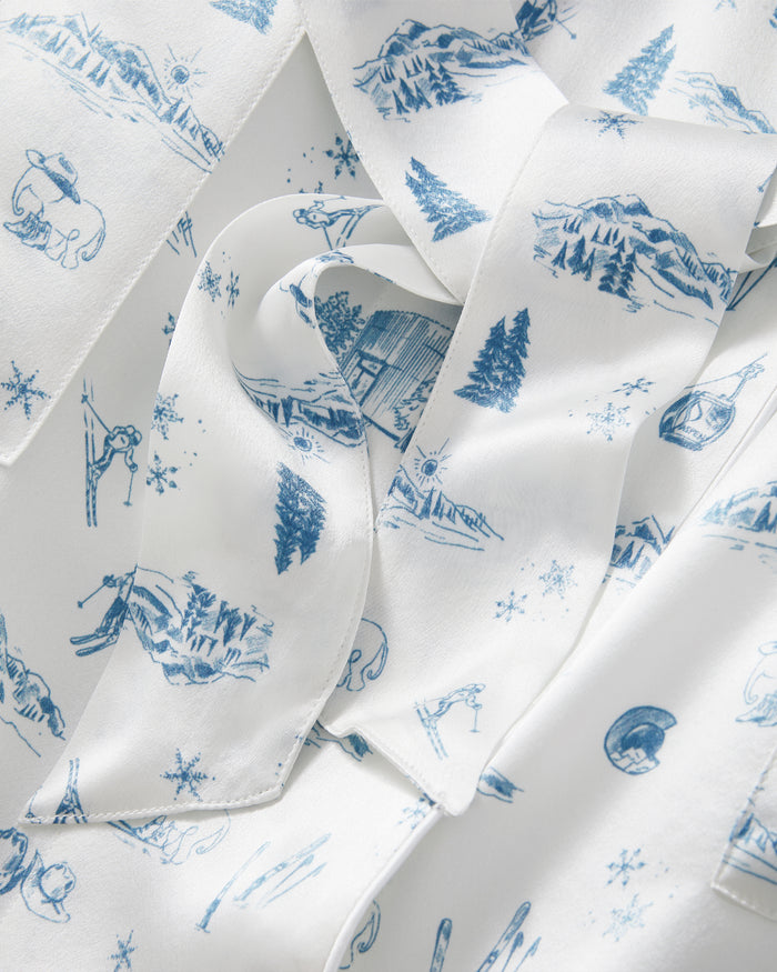 The Petite Plume x White Elephant Women’s Silk Robe features a white fabric adorned with blue illustrations of mountains, pine trees, cabins, skiers, snowflakes, and animals for a charming winter or alpine theme. The robe is shown softly folded to display the printed pattern from various angles.