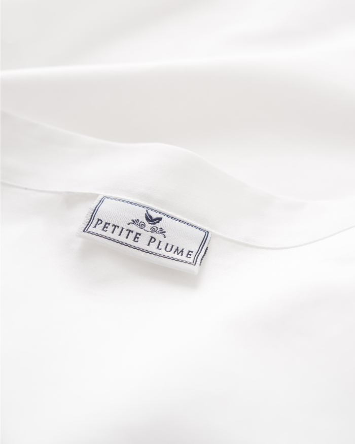 Close-up of a white sateen fabric label reading Petite Plume in blue, with decorative border and plume graphic, similar to those on Signature Luxe Sateen Bed Sheets by Petite Plume from luxury hotels.