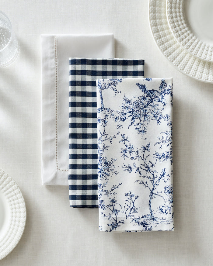 Three folded cotton napkins sit on a white tablecloth, featuring plain white, blue checkered, and Petite Plumes Signature Twill Napkin in Timeless Toile designs. Plates with textured rims adorn the festive tablescape alongside a partially visible glass.