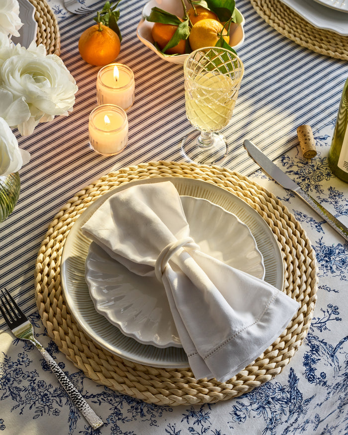 A festive tablescape includes white plates on a woven placemat, topped with Petite Plumes Signature Twill Napkins in White, accompanied by a wine glass and candles. Oranges with leaves rest on linens featuring blue floral patterns, surrounded by delicate white flowers.