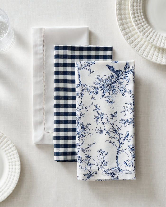 A set of three Petite Plume Signature Twill 100% cotton napkins—one plain white, one navy and white checkered, and one with a blue floral and bird design—add charm to the festive tablescape, arranged on a white tablecloth beside textured plates and a glass.
