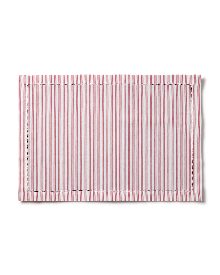 The table setting includes a white plate and red floral napkin, elevated by elegant linens and cream-handled utensils. Featuring Petite Plumes Signature Twill Placemats in Antique Red Ticking, it is adorned with white wine, meringues, a red ornament, and candle on the striped cloth.