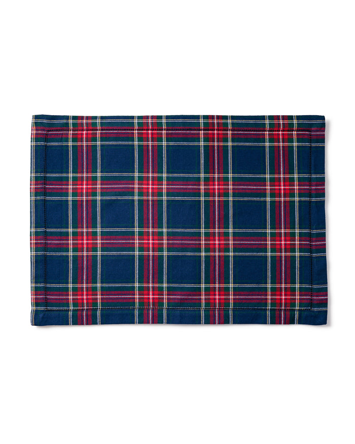 A cozy tablescape features white plates on Signature Twill Placemats in Windsor Tartan by Petite Plume, each with a matching napkin tied in a gold ring. A wine glass, fork, knife, corkscrew, candle, and vibrant bouquet of red flowers complete the look with versatile red, blue, and green hues.