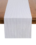 Petite Plumes Signature Twill Table Runner in Navy French Ticking, elegantly draped over a wooden table, adds a touch of classic style with its 100% cotton blue and white vertical stripes, perfect for enhancing festive tablescapes.