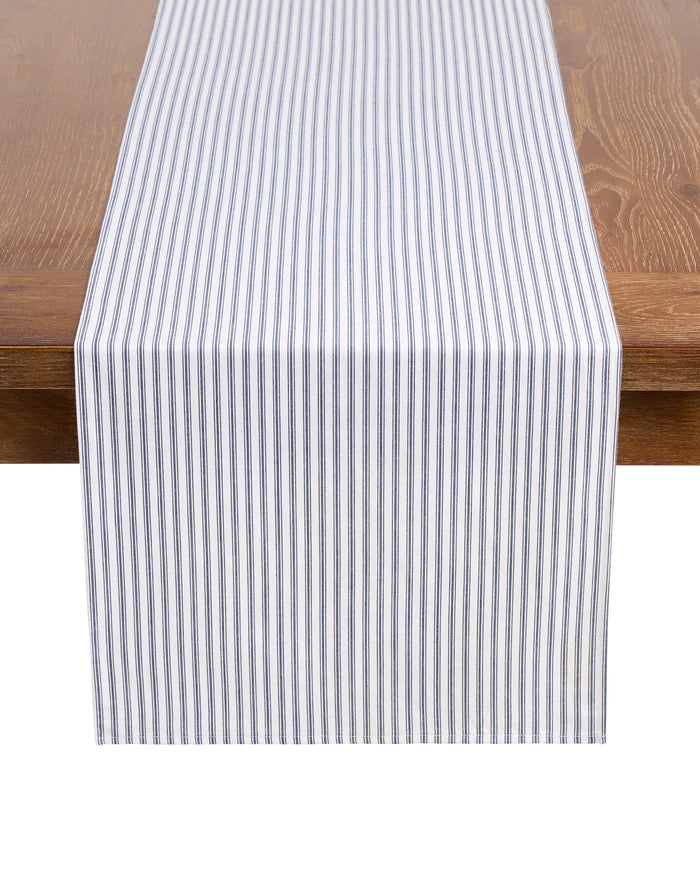 Petite Plumes Signature Twill Table Runner in Navy French Ticking, elegantly draped over a wooden table, adds a touch of classic style with its 100% cotton blue and white vertical stripes, perfect for enhancing festive tablescapes.