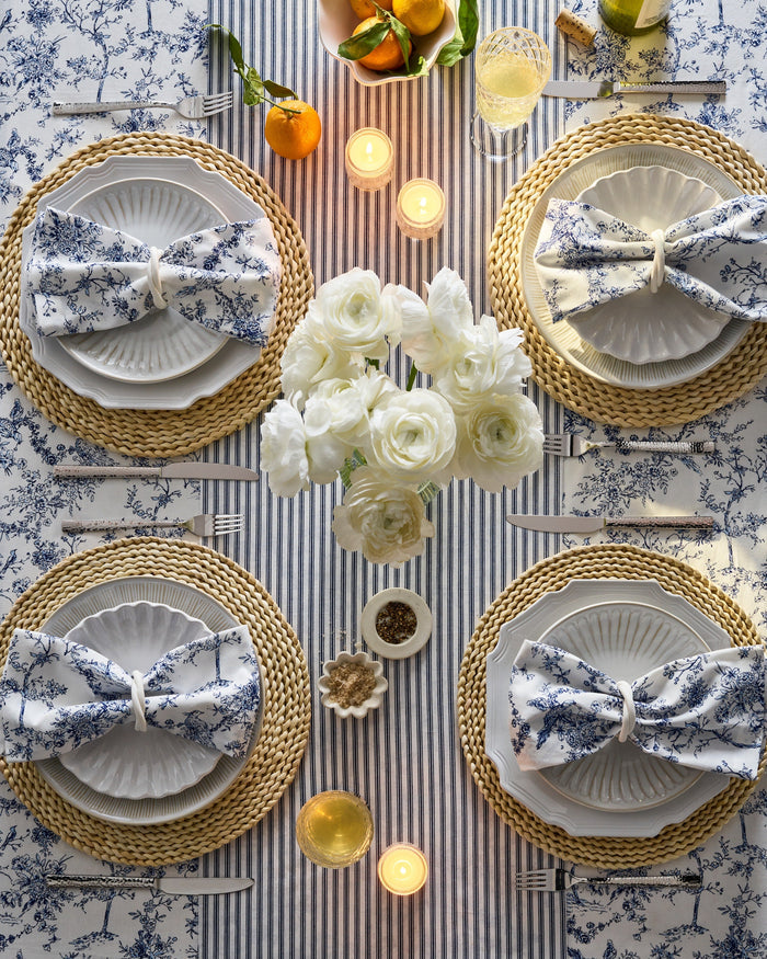 Top view of a chic dining table set for four with white plates on woven placemats, floral napkins, a festive tablescape featuring a white flower centerpiece, candles, drinks, and oranges on Petite Plumes Signature Twill Table Runner in Navy French Ticking.