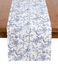 A wooden table is elegantly adorned with 100% cotton table linens, highlighted by Petite Plumes Signature Twill Table Runner in Timeless Toile, showcasing beautiful blue florals and birds.