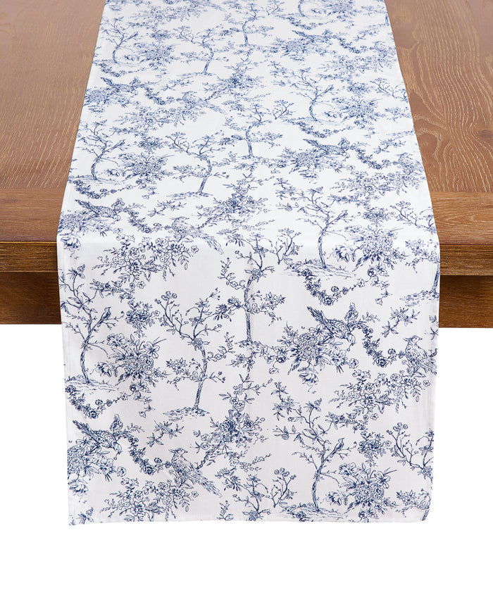 A wooden table is elegantly adorned with 100% cotton table linens, highlighted by Petite Plumes Signature Twill Table Runner in Timeless Toile, showcasing beautiful blue florals and birds.