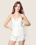 A woman models the Petite Plume Womens Silk Lace Cami Short Set in White, showcasing a satin camisole and matching shorts with floral lace trim. She stands against a plain beige background, hands on her hips and a neutral expression.