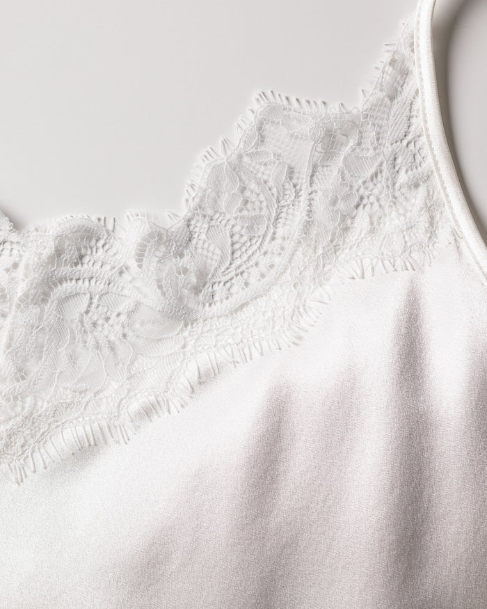 A close-up of the intricate white floral lace trim from Petite Plumes Womens Silk Lace Cami Short Set on a satin-like surface. The elegant design hints at luxury silk sleepwear, with a plain, light background enhancing its sophistication.
