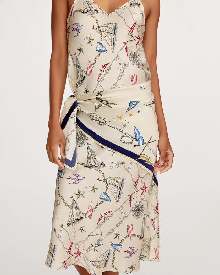 A woman wearing the Silk Scarf in Regatta Royale, featuring a cream base with nautical-themed prints such as anchors, ropes, compasses, and sailboats. The scarf is styled with a blue-trimmed wrap detail at the waist. Her head is not visible.