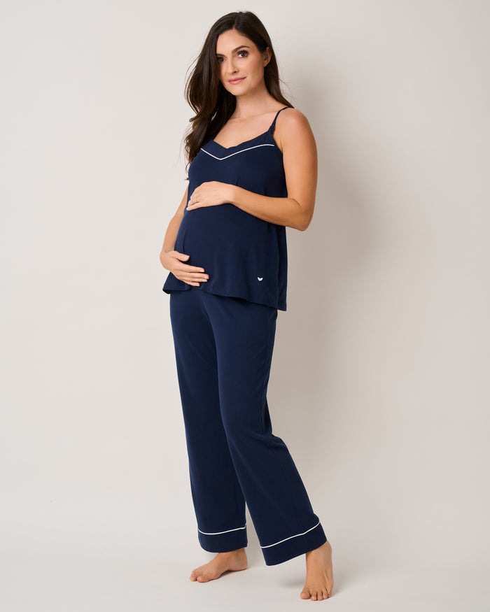 A pregnant woman, gently cradling her belly, stands barefoot in a Petite Plume Basics Maternity Set in Navy. Her Pima cotton camisole and matching pajama pants with white piping blend seamlessly against the simple, light-colored wall.