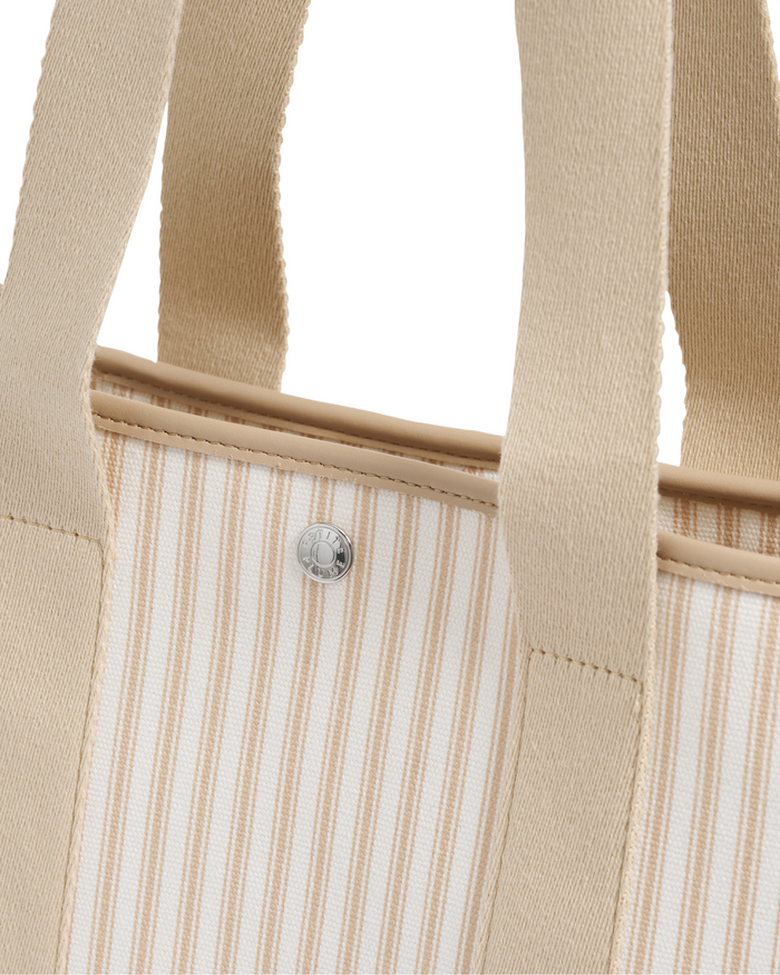 A close-up of The Beckett Canvas Tote Bag in Camel Ticking, featuring beige and white stripes, thick beige handles, and a silver button detail near the top edge.