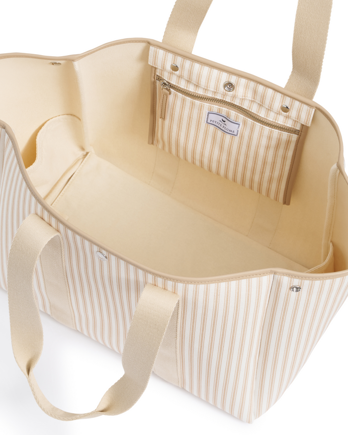 The Beckett Canvas Tote Bag in Camel Ticking features a large, open design with beige and white vertical stripes, complementary beige handles, and a practical inner zippered pocket attached to the lining. Its spacious interior remains unlined, showcasing neutral tones throughout.