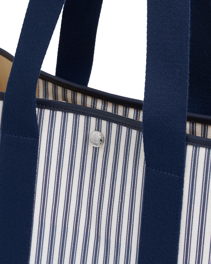 Close-up of The Beckett Canvas Tote Bag in Navy French Ticking, featuring navy handles and blue vertical stripes on a white background. A silver button is visible near the top edge.