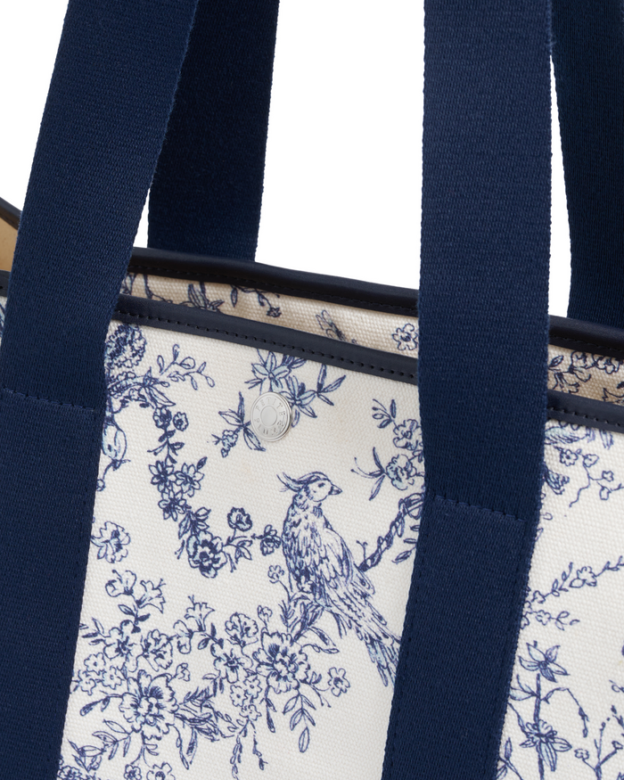 Close-up of The Beckett Canvas Tote Bag in Timeless Toile, featuring wide navy blue handles and a classic blue and white floral toile pattern with a bird nestled among the branches. A small round metal emblem is affixed near the top edge.