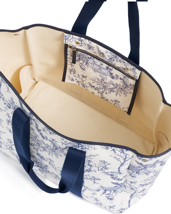 The Beckett Canvas Tote Bag in Timeless Toile is a spacious, open tote featuring a navy and cream floral print, sturdy navy handles, and an interior pocket with a coordinating pattern and zipper closure. The bag is pictured empty and viewed from above.