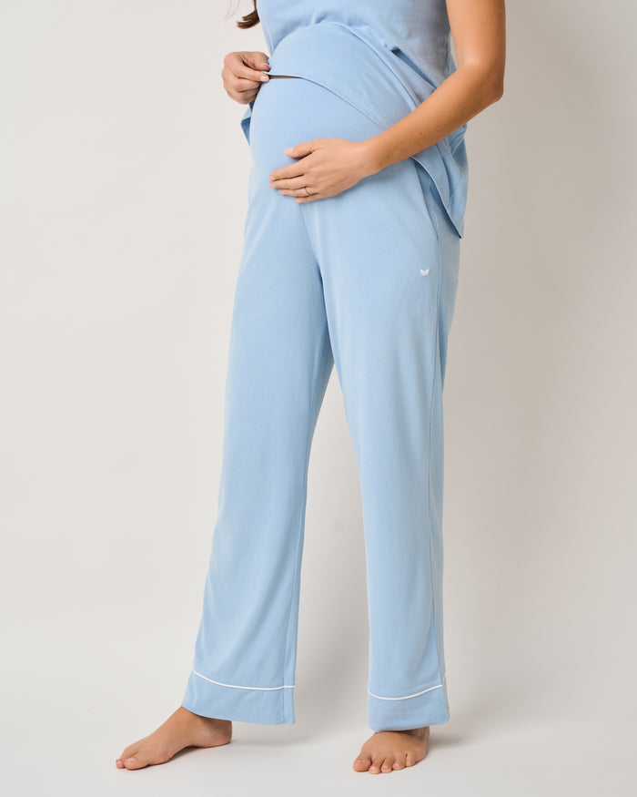 A pregnant person stands against a plain background, wearing The Cozy Maternity Set in Periwinkle by Petite Plume. They cradle their belly with both hands, exuding tranquility, as one foot is slightly raised to rest on the toes.