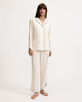 A person stands barefoot on a white background, wearing The Margaux Lightweight Pima Pajama Set in Ivory, featuring long sleeves, buttons, and pants with dark piping along the edges.