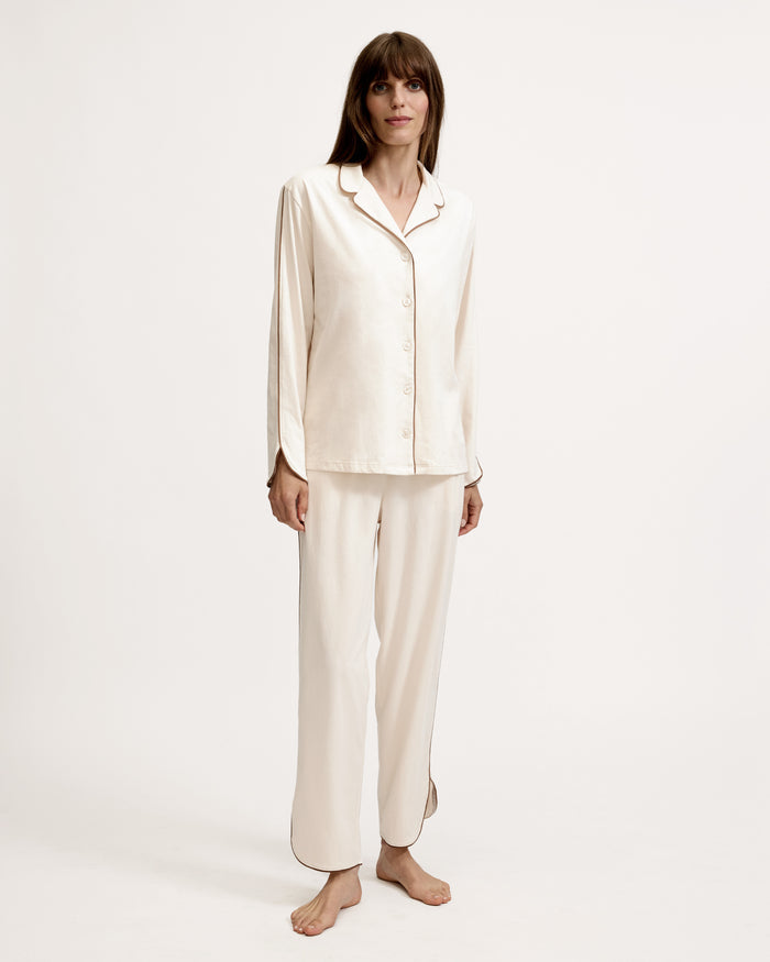 A person stands barefoot on a white background, wearing The Margaux Lightweight Pima Pajama Set in Ivory, featuring long sleeves, buttons, and pants with dark piping along the edges.