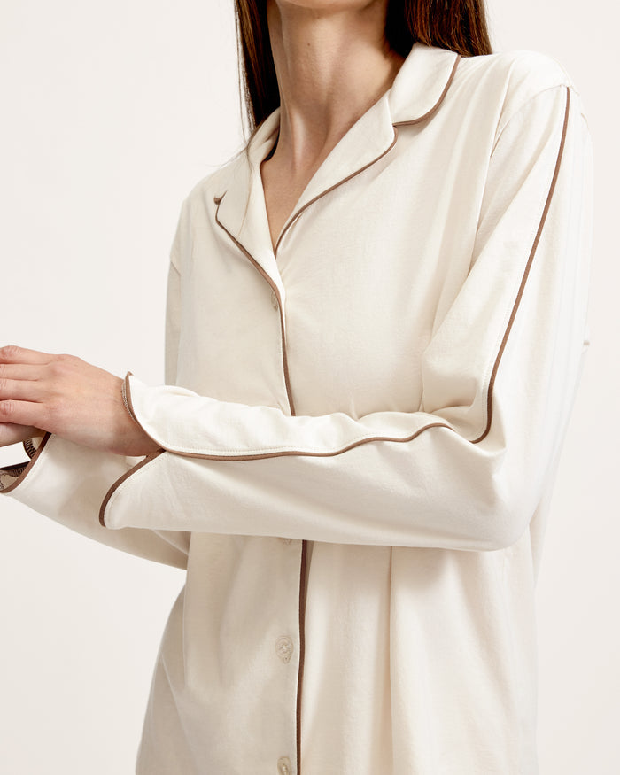 A person stands barefoot on a white background, wearing The Margaux Lightweight Pima Pajama Set in Ivory, featuring long sleeves, buttons, and pants with dark piping along the edges.