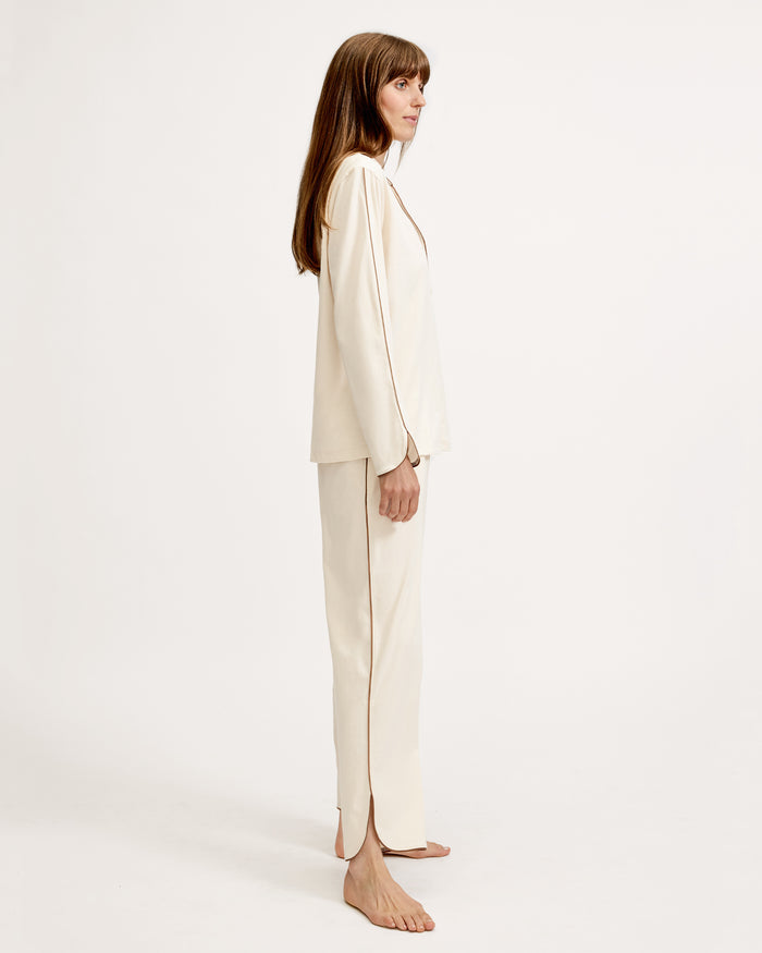 A person stands barefoot on a white background, wearing The Margaux Lightweight Pima Pajama Set in Ivory, featuring long sleeves, buttons, and pants with dark piping along the edges.