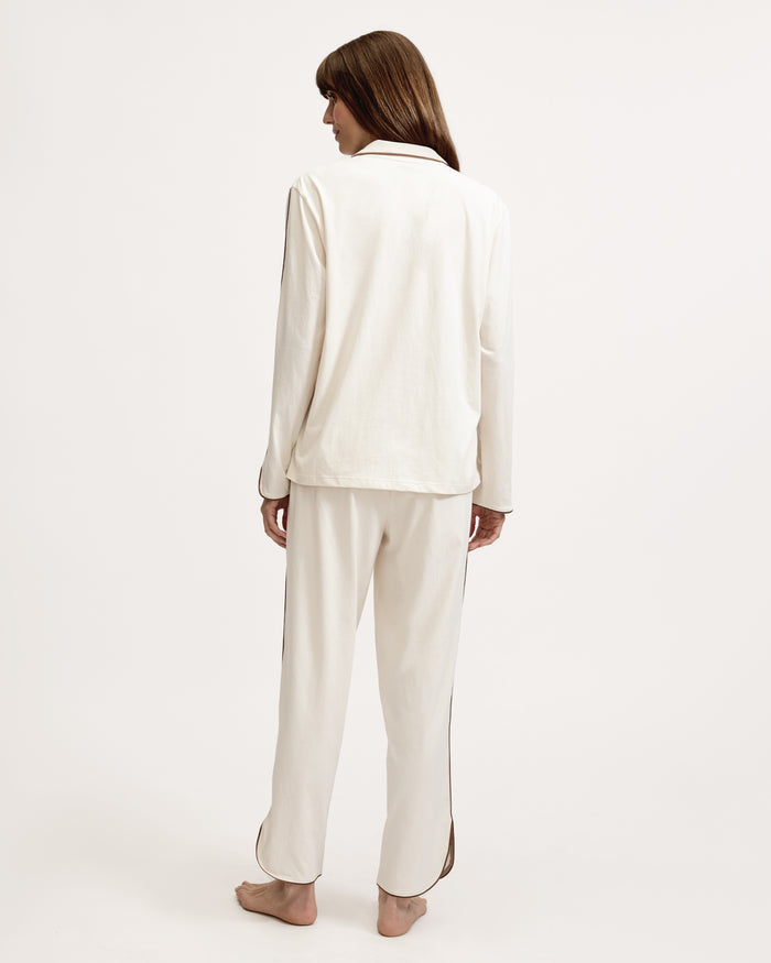 A person stands barefoot on a white background, wearing The Margaux Lightweight Pima Pajama Set in Ivory, featuring long sleeves, buttons, and pants with dark piping along the edges.
