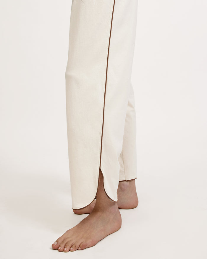 A person stands barefoot on a white background, wearing The Margaux Lightweight Pima Pajama Set in Ivory, featuring long sleeves, buttons, and pants with dark piping along the edges.