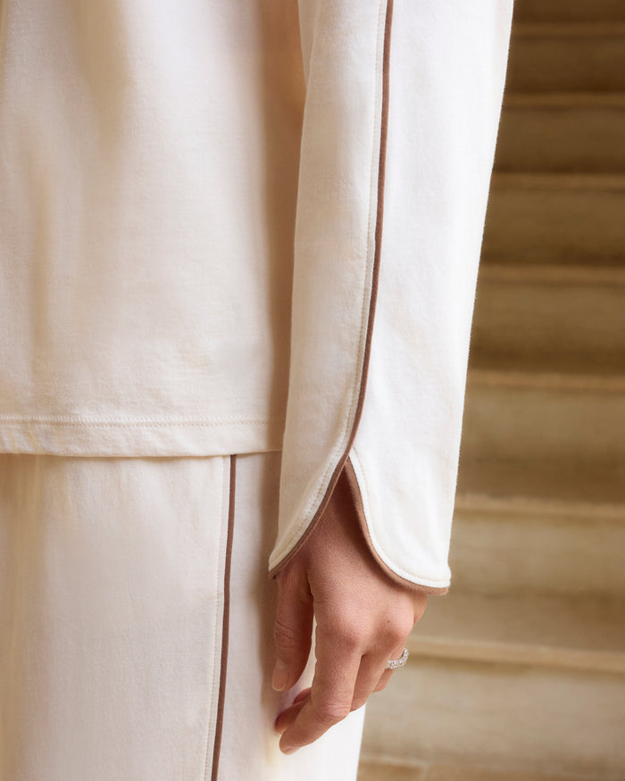 A person stands barefoot on a white background, wearing The Margaux Lightweight Pima Pajama Set in Ivory, featuring long sleeves, buttons, and pants with dark piping along the edges.