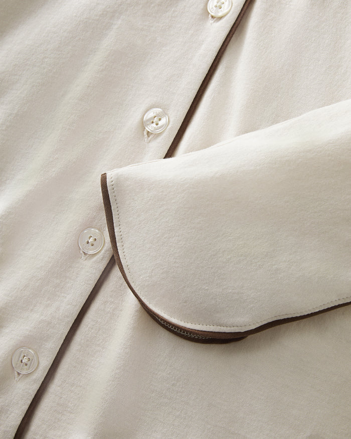 A person stands barefoot on a white background, wearing The Margaux Lightweight Pima Pajama Set in Ivory, featuring long sleeves, buttons, and pants with dark piping along the edges.