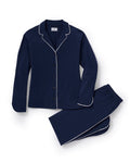 A person stands barefoot in The Margaux Pima Pajama Set in Navy, featuring white piping, a buttoned long-sleeve top, and loose-fitting pants. The background is plain and light-colored.
