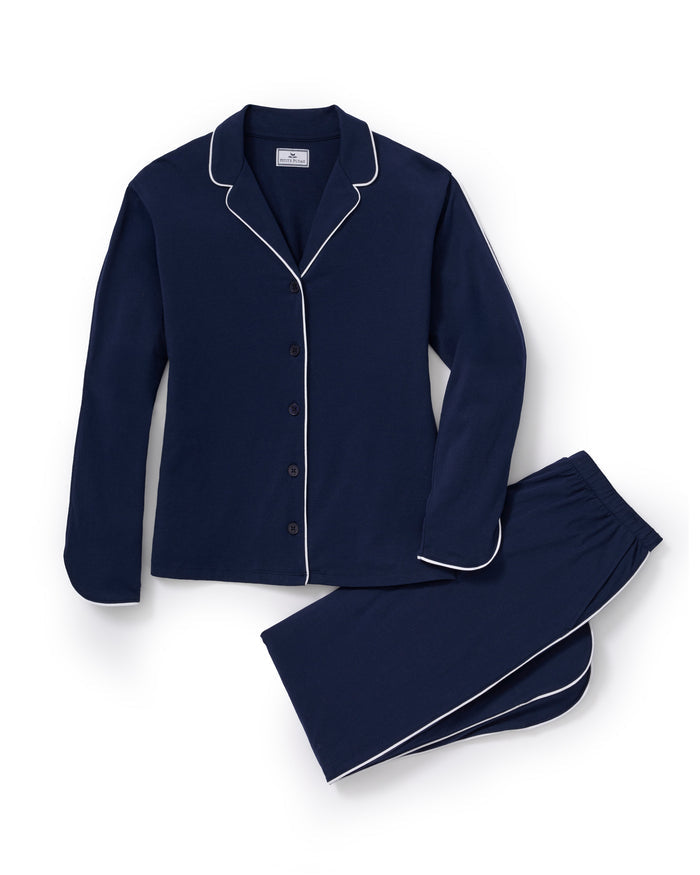 A person stands barefoot in The Margaux Pima Pajama Set in Navy, featuring white piping, a buttoned long-sleeve top, and loose-fitting pants. The background is plain and light-colored.
