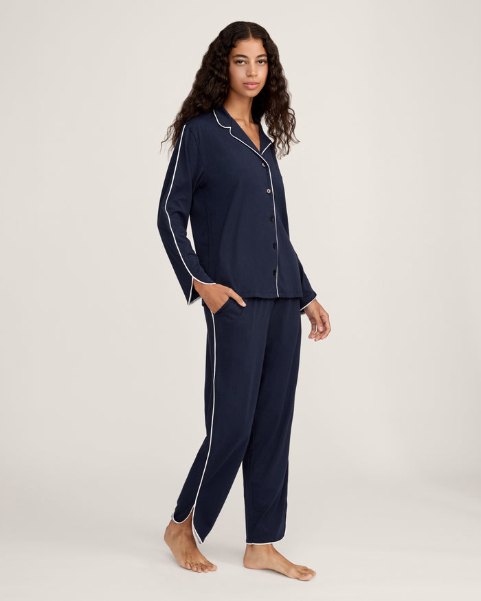 A person stands barefoot in The Margaux Pima Pajama Set in Navy, featuring white piping, a buttoned long-sleeve top, and loose-fitting pants. The background is plain and light-colored.