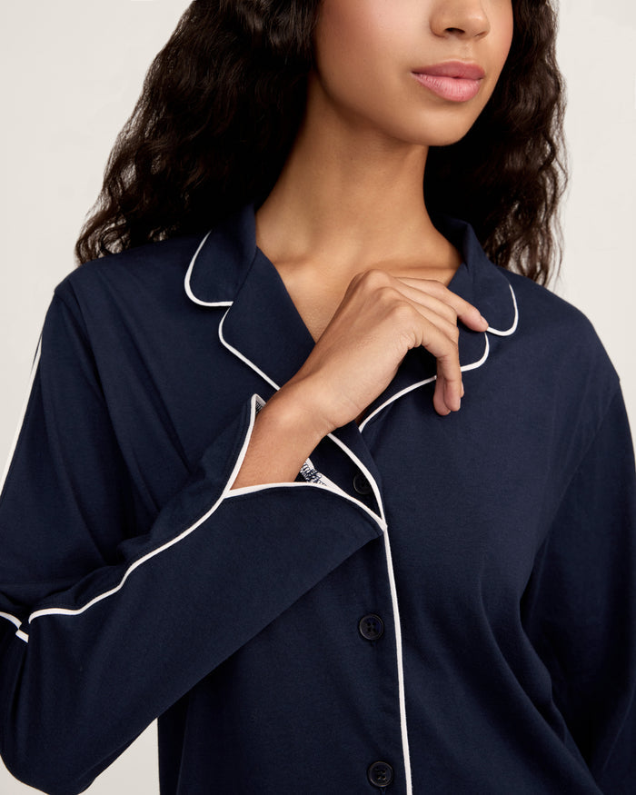A person stands barefoot in The Margaux Pima Pajama Set in Navy, featuring white piping, a buttoned long-sleeve top, and loose-fitting pants. The background is plain and light-colored.