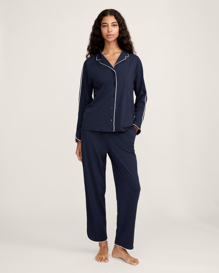 A person stands barefoot in The Margaux Pima Pajama Set in Navy, featuring white piping, a buttoned long-sleeve top, and loose-fitting pants. The background is plain and light-colored.