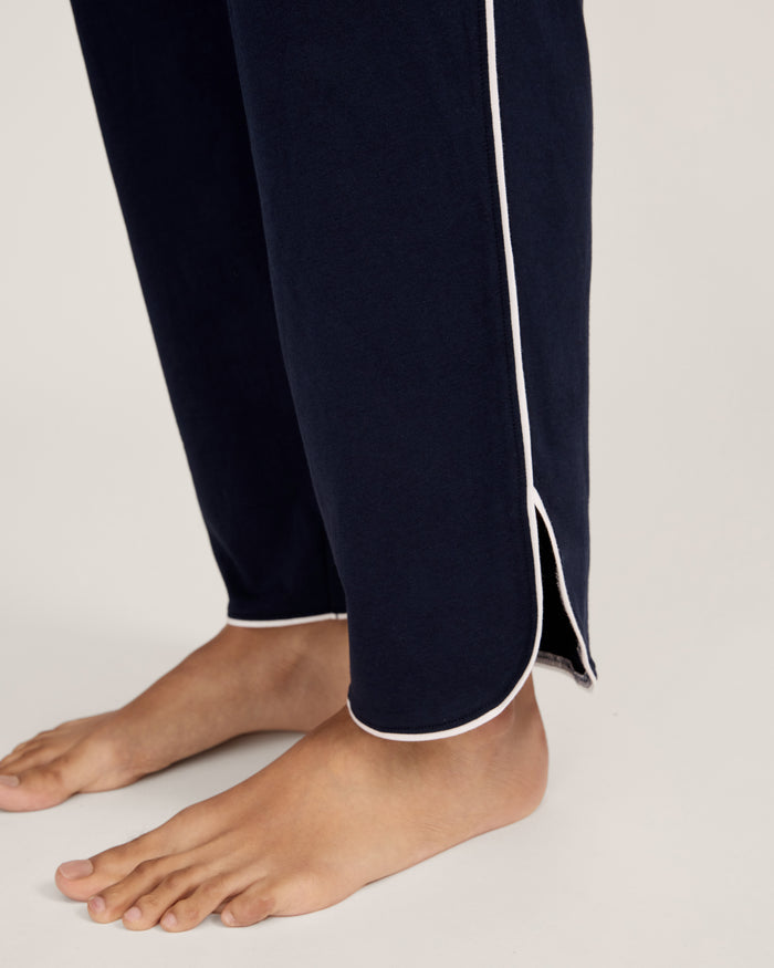 A person stands barefoot in The Margaux Pima Pajama Set in Navy, featuring white piping, a buttoned long-sleeve top, and loose-fitting pants. The background is plain and light-colored.