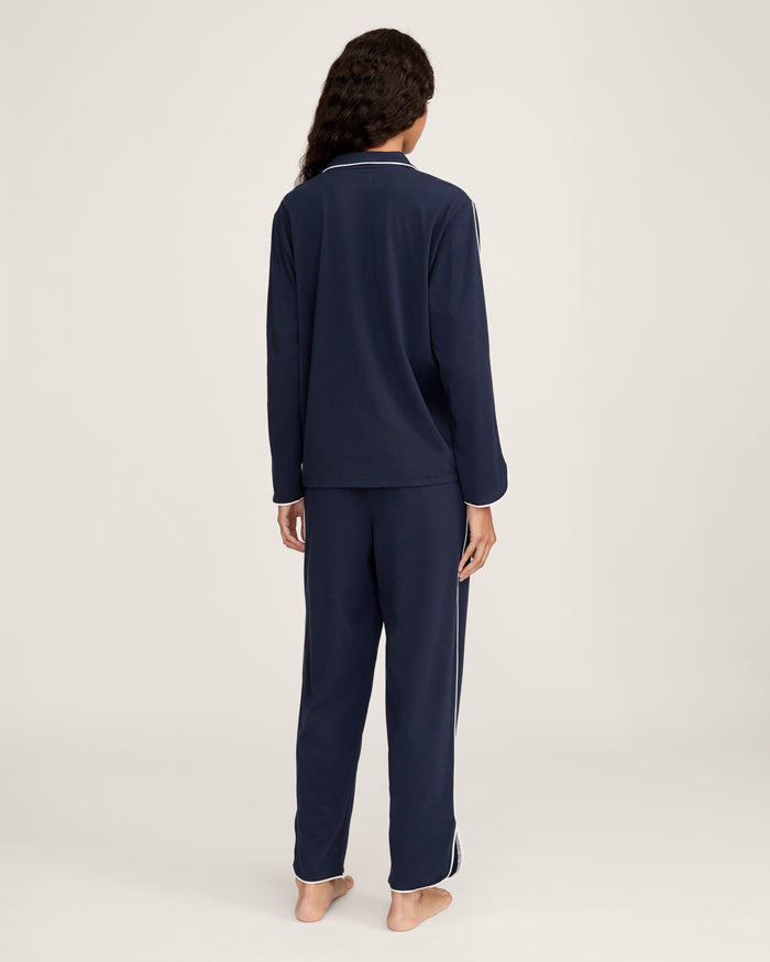 A person stands barefoot in The Margaux Pima Pajama Set in Navy, featuring white piping, a buttoned long-sleeve top, and loose-fitting pants. The background is plain and light-colored.