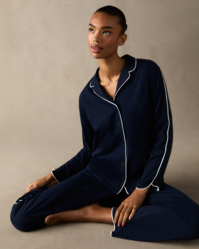 A person stands barefoot in The Margaux Pima Pajama Set in Navy, featuring white piping, a buttoned long-sleeve top, and loose-fitting pants. The background is plain and light-colored.