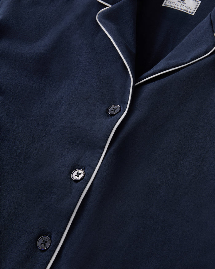 A person stands barefoot in The Margaux Pima Pajama Set in Navy, featuring white piping, a buttoned long-sleeve top, and loose-fitting pants. The background is plain and light-colored.