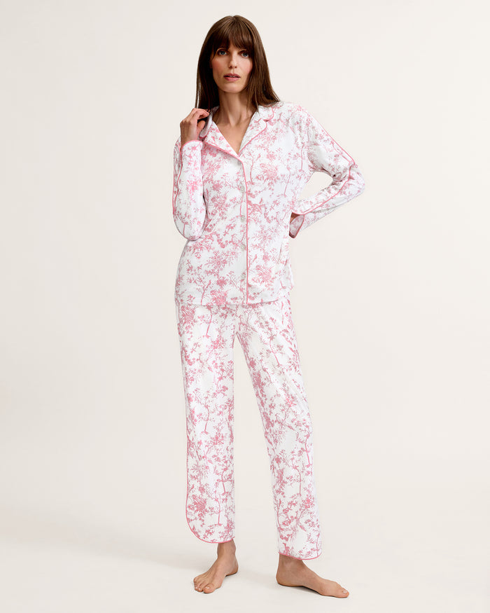 A woman stands barefoot on a plain background, wearing The Margaux Pima Pajama Set in Pink Timeless Toile. She has straight brown hair and lightly touches her chin with one hand.