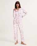 A woman stands barefoot on a plain background, wearing The Margaux Pima Pajama Set in Pink Timeless Toile. She has straight brown hair and lightly touches her chin with one hand.