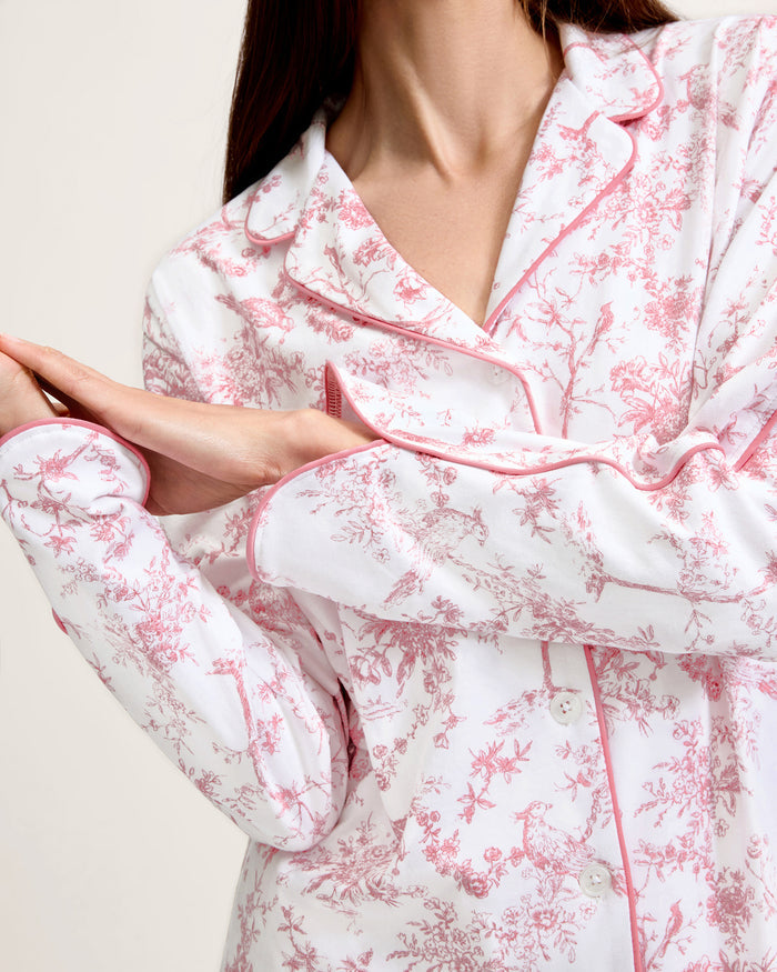 A woman stands barefoot on a plain background, wearing The Margaux Pima Pajama Set in Pink Timeless Toile. She has straight brown hair and lightly touches her chin with one hand.