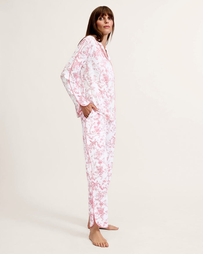 A woman stands barefoot on a plain background, wearing The Margaux Pima Pajama Set in Pink Timeless Toile. She has straight brown hair and lightly touches her chin with one hand.