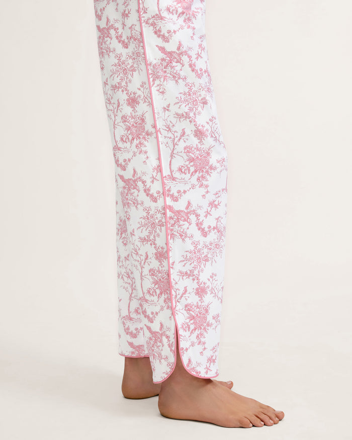 A woman stands barefoot on a plain background, wearing The Margaux Pima Pajama Set in Pink Timeless Toile. She has straight brown hair and lightly touches her chin with one hand.
